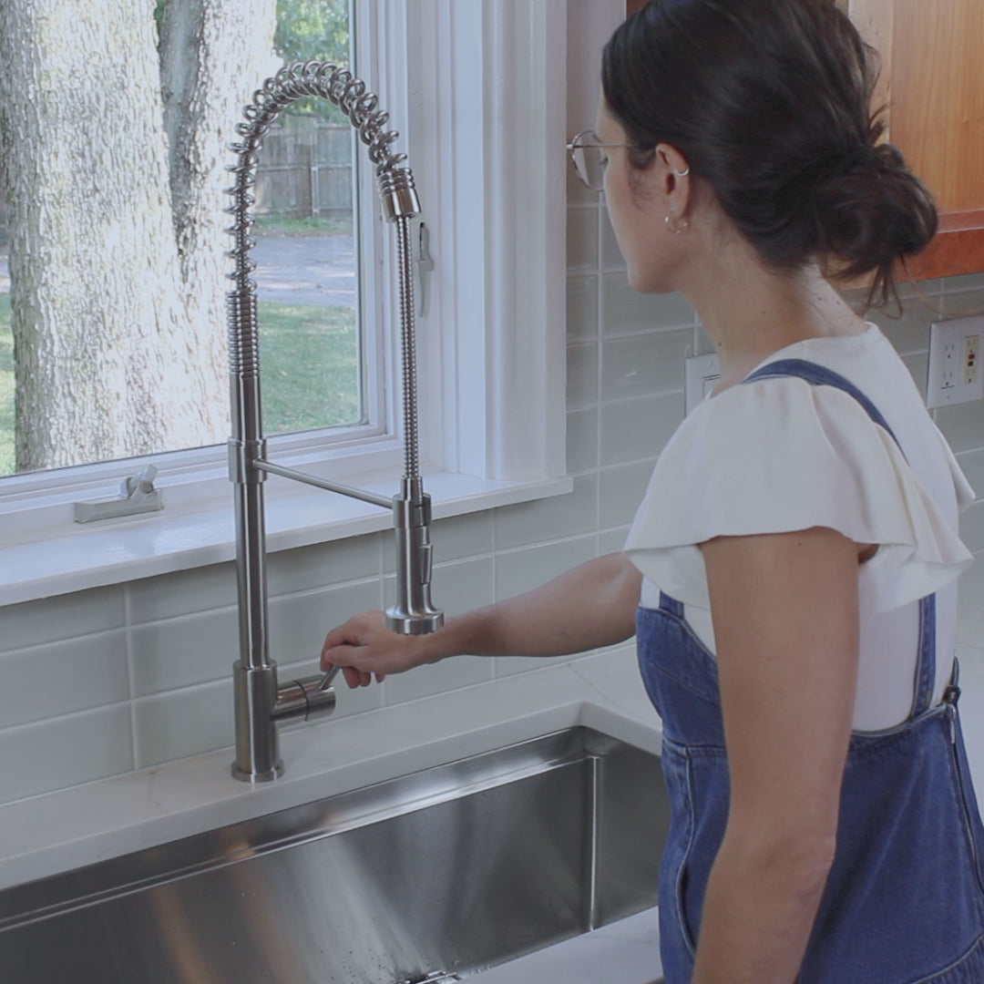 Woman using the Jerry stainless steel kitchen faucet, adjusting the handle while standing in a modern kitchen with a large window view.