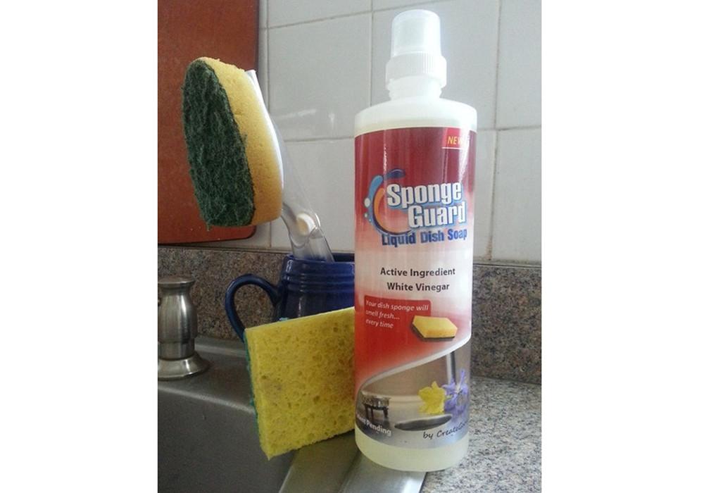 Sponge-Guard Liquid Dish Soap bottle placed near a kitchen sink with a sponge and dish brush, emphasizing its cleaning and sponge-protecting properties.