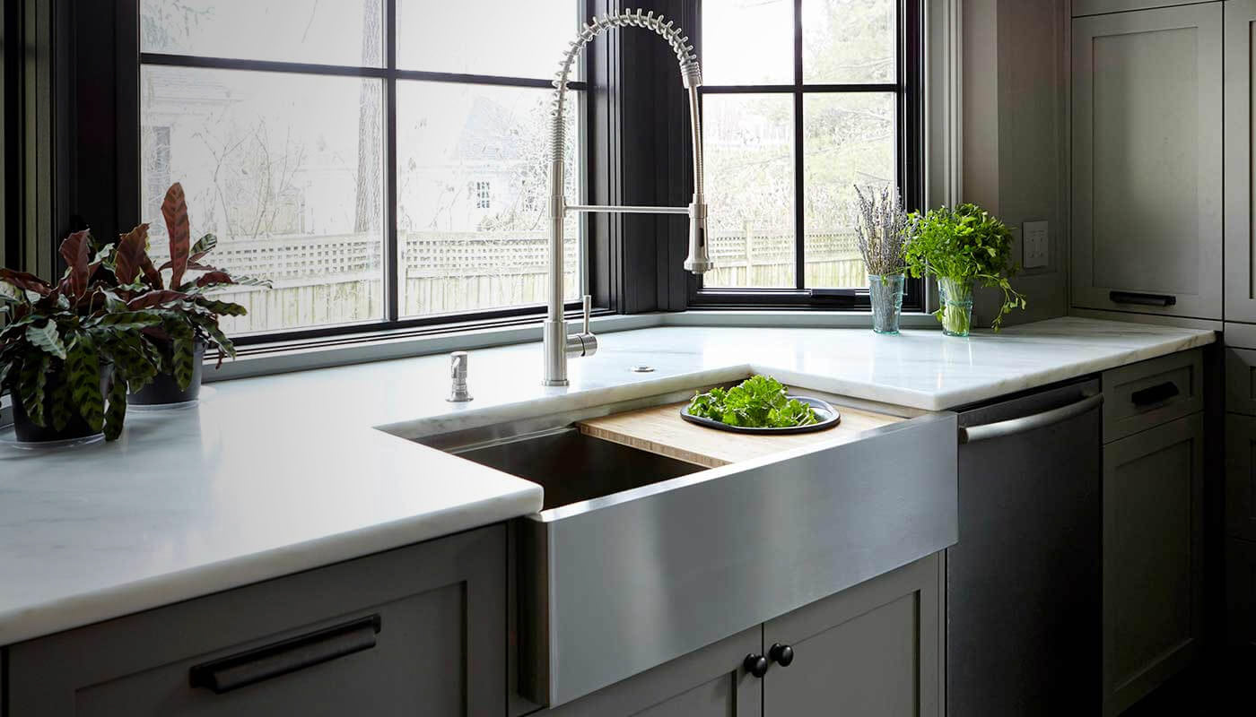 Stainless steel apron front farmhouse workstation sink in a marble kitchen with bamboo cutting board and colander on the ledge
