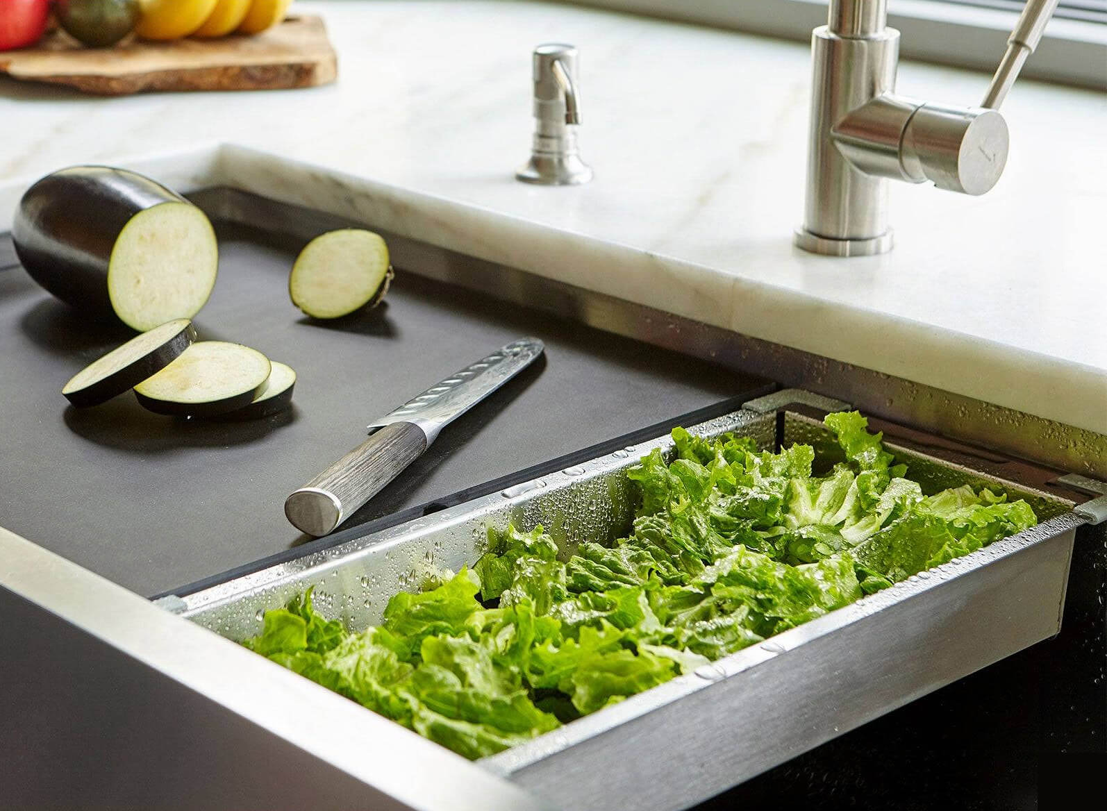 Washed lettuce draining in a stainless steel prep basin inside a workstation sink with sliced eggplant on the cutting board