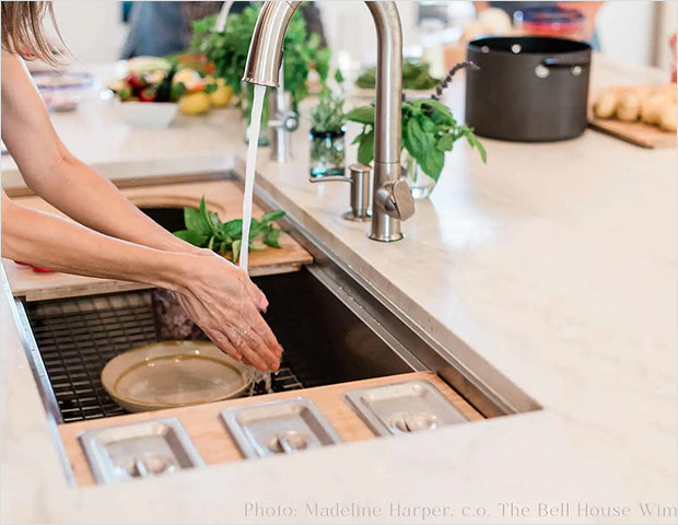 Hands rinsing fresh basil in a large stainless steel workstation sink with sink grid, prep pans and cutting board on the ledge