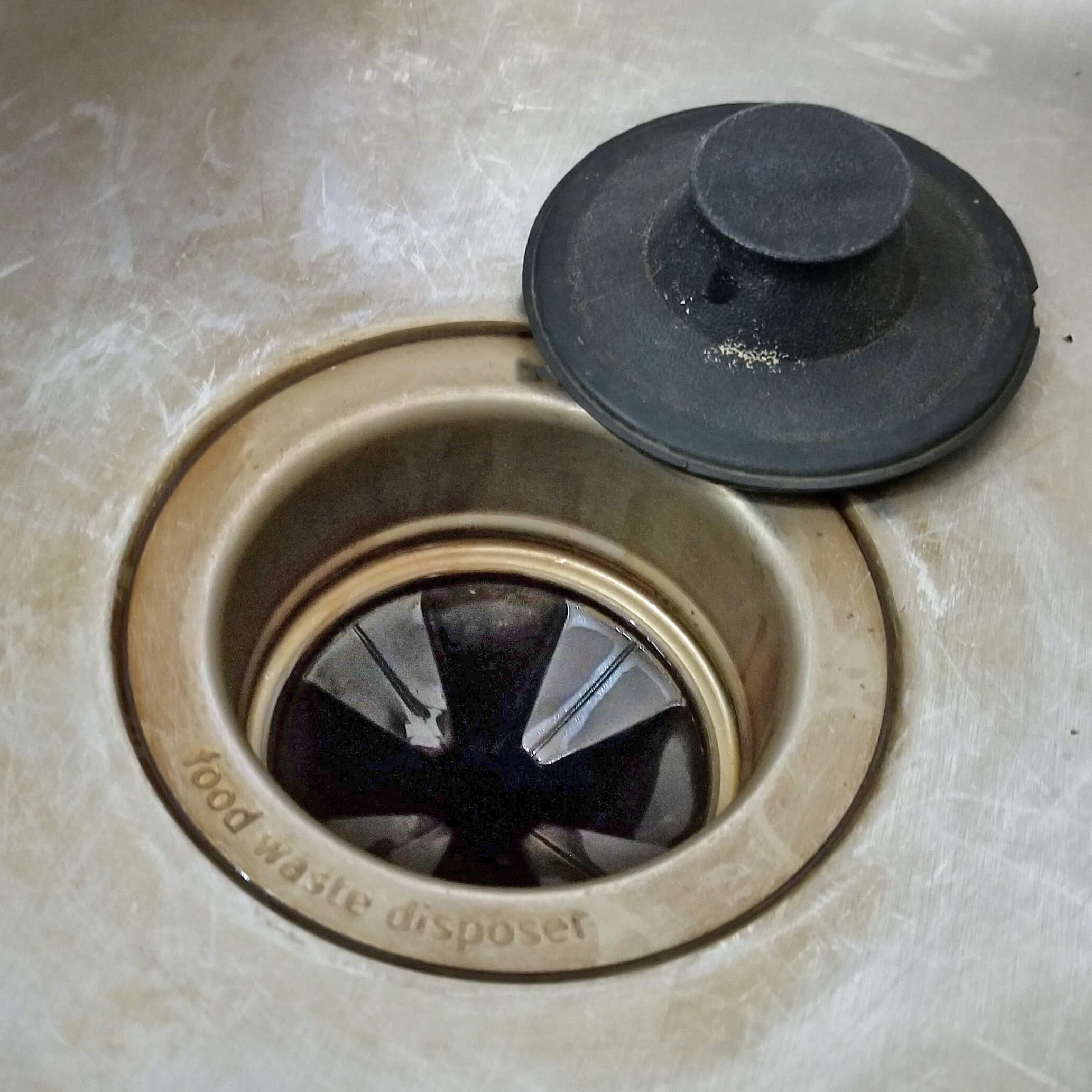 Grimy conventional garbage disposal flange with worn rubber splash guard and corroded seam