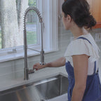 Woman using the Jerry stainless steel kitchen faucet, adjusting the handle while standing in a modern kitchen with a large window view.