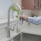 Bella stainless steel kitchen sink faucet in use, with a person operating the pull-down spray head over a modern sink setup.