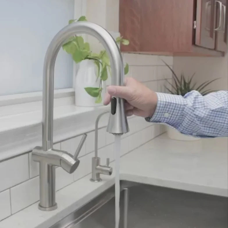 Bella stainless steel kitchen sink faucet in use, with a person operating the pull-down spray head over a modern sink setup.