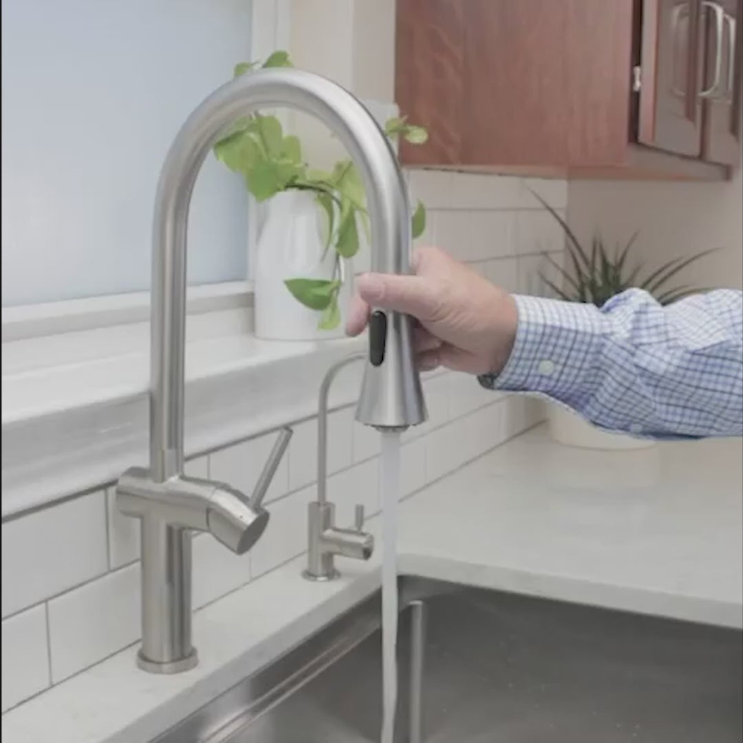 Bella stainless steel kitchen sink faucet in use, with a person operating the pull-down spray head over a modern sink setup.